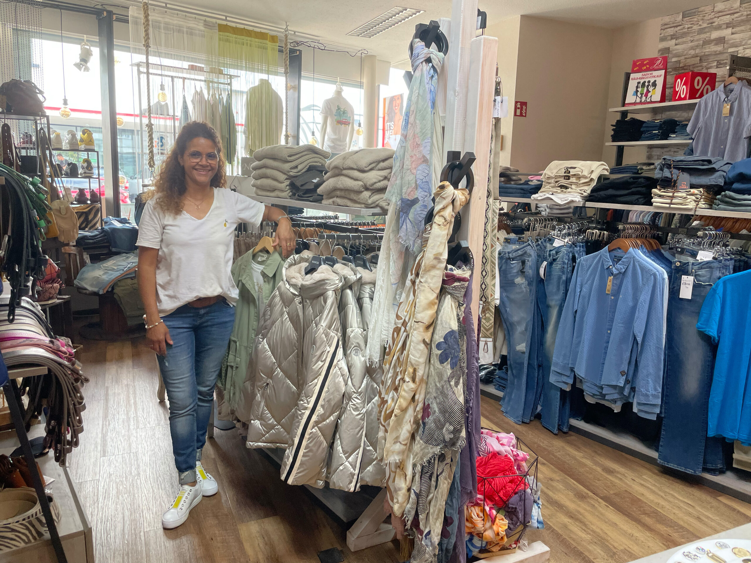 Bild zeigt die Inhaberin von Just Once Fashion in Marbach, Daniela Zäch in der Mitte des Ladens. Sie trägt eine Jeans und ein weißes T-Shirt und lehnt an einem Kleiderständer mit Herbstmoden. Die Haare trägt sie halb offen.